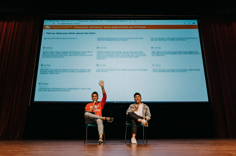 Two men seated on stage, one raising his hand, with a large projected presentation screen behind them.